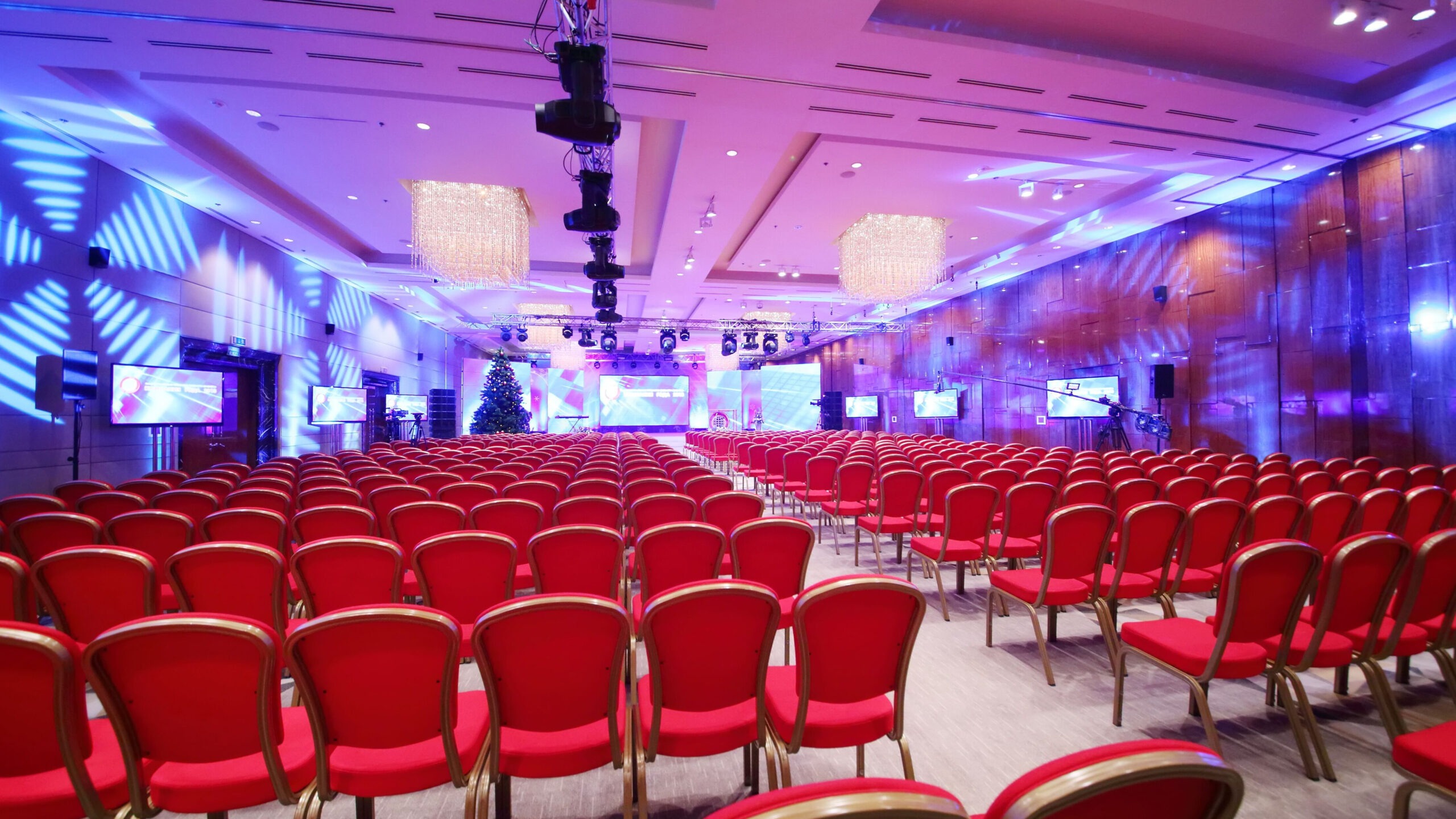A spacious room featuring red chairs arranged for an audience, with a stage set up at the front for presentations or performances.